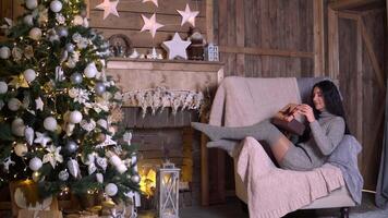 familj jul, roligt fest, stanna kvar på Hem, ny år firande. kvinna i grå Tröja och strumpor öppnas en gåva låda medan Sammanträde i en stol på jul eve video
