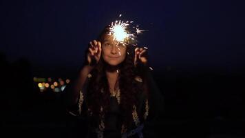 joven hermosa mujer con largo oscuro pelo sostiene fuegos artificiales a noche en el antecedentes de el ciudad y se regocija lento movimiento. video