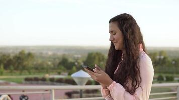 un' bellissimo ragazza con lungo capelli passeggiate nel il parco vicino il Ferris ruota e usi un' smartphone. 4k. video