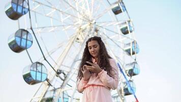 uma lindo menina dentro uma vestir usa uma Smartphone em pé perto a ferris roda 0,4k video