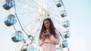 un hermosa niña en un vestir usos un teléfono inteligente en pie cerca el ferris rueda .4k video