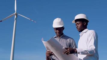 Two male African American electrical engineers stand against the backdrop of a windmill at an air power plant with a drawing in their hands and read them video