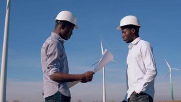 Two male African American electrical engineers stand against the backdrop of a windmill at an air power plant. They hold the drawing in their hands and examine it video