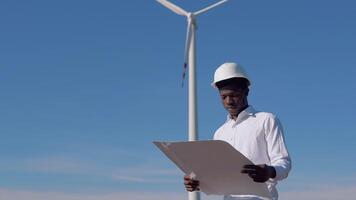 African American electrician engineer in a helmet stands against the backdrop of a windmill at an air power plant and reads a drawing. Wind power turbines generating clean renewable energy video