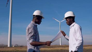 Two male African American electrical engineers stand against the backdrop of a windmill at an air power plant. They hold the drawing in their hands and examine it video