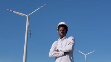 africano americano electricista ingeniero soportes en el antecedentes de un molino a un aire poder planta y muestra un pulgar arriba. viento poder turbinas generando limpiar renovable energía video