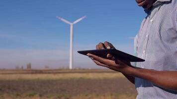 An African-American electrician in a helmet stands against the backdrop of a windmill at an air power plant and looks at a tablet and swipes across the screen. Close-up view of hands and tablet video