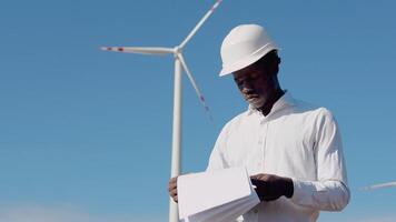 African american electrical engineer standing on the background of a windmill at an air power plant reading records video