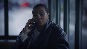 A woman in eyewear sits by the window, chatting on her cell phone video