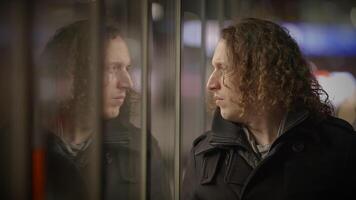 In this video, a man with long curly hair stands in front of a glass door and window. He is seen looking out of the window at night, creating a mysterious and contemplative atmosphere video