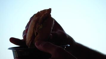 silhueta do uma homem desfrutando comendo do uma Hamburger. lado Visão retrato do uma pessoa saboreando uma delicioso, satisfatório hamburguer com ótimo apetite. velozes Comida indulgência. video