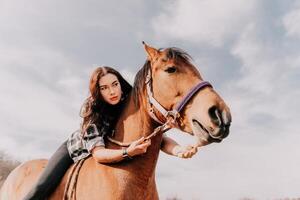 Young happy woman in hat with her horse in evening sunset light. Outdoor photography with fashion model girl. Lifestyle mood. Concept of outdoor riding, sports and recreation. photo