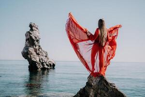 Woman travel sea. Young Happy woman in a long red dress posing on a beach near the sea on background of volcanic rocks, like in Iceland, sharing travel adventure journey photo