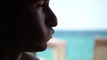 Young, happy man eats sandwich with cheese, sausage and ketchup on beach by sea. Takeaway food. Lunch break. Silhouette head of male tourist sitting on beach and eating sandwich, slow-motion. Profile video