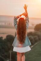 contento mujer en pie con su espalda en el puesta de sol en naturaleza en verano con abierto manos. romántico hermosa novia en blanco boho vestir posando con montañas en puesta de sol foto