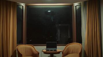 Nighttime office with two chairs and a laptop on a desk, framed by curtains and a city view through a large window. video