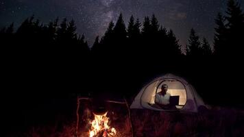 Man Works behind a Laptop in a tent near a fire at night. Timelapse of moving stars and The Milky Way in night sky over the forest and tent. Travel concept 4K video