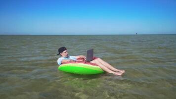 A young business person is enthusiastically working behind a laptop swaying on the waves of sea sitting on a inflatable circle. 4K video