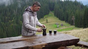 A man pours coffee from a coffee maker in the morning in the mountains. Wooden houses of shepherds in the background. Travel concept video