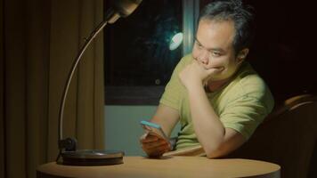 Stressed man sitting at a table under lamp light with a phone, looking tired and frustrated. video