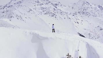 Success concept. A snowboarder in winter attire stands on a mild slope in Verbier, Switzerland, overlooking snow capped mountains and pines. A man standing on a stone cliff over the clouds. video