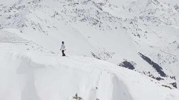 Success concept. A snowboarder in winter attire stands on a mild slope in Verbier, Switzerland, overlooking snow capped mountains and pines. A man standing on a stone cliff over the clouds. video