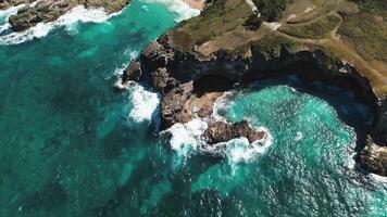 Bird's-eye perspective of breathtaking rocky coastline being hit by waves against cliffs under cloudless, sunny sky, with water so transparent it resembles turquoise. Flight forward video