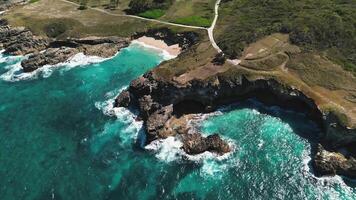 Breathtaking aerial view captures rocky coastline being lashed by waves against towering cliffs under clear, sunny sky. Water is so clear and turquoise that it almost looks transparent. Dominican video