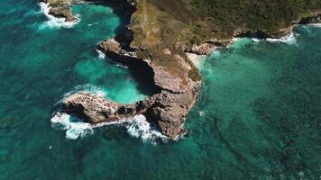 aérien vue de étourdissant rocheux littoral avec vagues martèlement falaises en dessous de clair, ensoleillé ciel, avec l'eau donc clair c'est turquoise. drone est en mouvement le long de une circulaire trajectoire. video