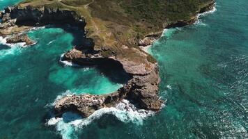 Aerial view of waves crash against rocks in turquoise Caribbean Sea near Macao, Dominican Republic, creating swirling patterns along rocky, picturesque coastline of tropical island, ocean. Zoom in video