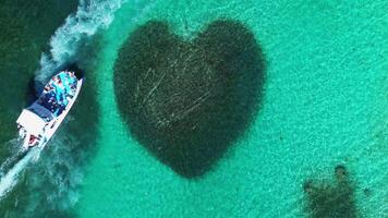 Aerial top view captures heart-shaped coral reef embraced by vibrant turquoise waters in tropical paradise located in Caribbean Sea. Boat carrying tourists sails past. video