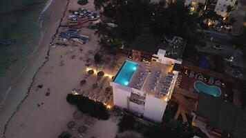 aéreo Noche cinematográfico ver sigue orbita trayectoria, enfoque en edificio con techo piscina con vista a Oceano línea costera. orilla punteado con playa sillas y barcos paisaje urbano luces antecedentes video