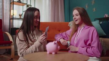 Girls friends siblings sitting on floor and take turns dropping dollar banknote into piggy bank video