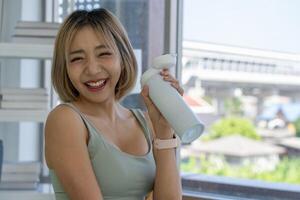 retrato de joven asiático deportivo sonriente mujer Bebiendo agua después rutina de ejercicio a hogar foto
