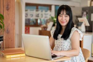 A cute Asian woman sits at a table in a coffee shop with her laptop computer, smiling at the camera. photo