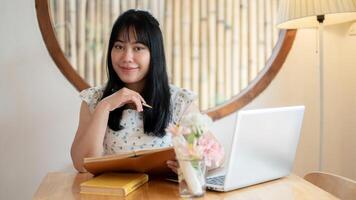 A cute Asian woman sits at a table in a coffee shop with her laptop computer, smiling at the camera. photo
