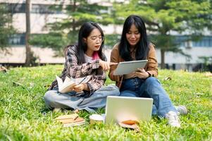 dos joven estudiantes son sentado en el césped en un parque, que se discute alguna cosa en un digital tableta. foto