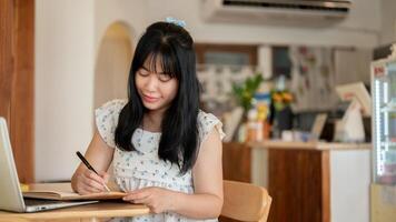 un hermosa asiático mujer enfoque en escritura alguna cosa en un cuaderno mientras sentado en un café tienda. foto