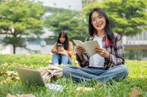 A smiling Asian female student is reading a book while sitting on the grass in a campus park. photo