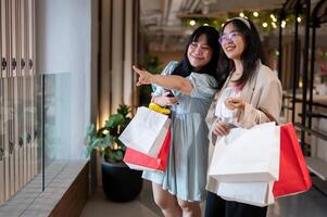 Two happy young Asian women pointing and smiling in a stylish shopping area, holding shopping bags. photo