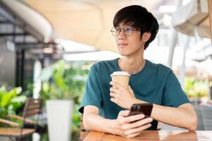 un hombre participación un café taza y un teléfono inteligente, mirando lejos, sentado a un al aire libre mesa de un cafetería. foto