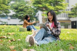 un hembra Universidad estudiante es estudiando en su digital tableta mientras sentado en césped en un parque. foto