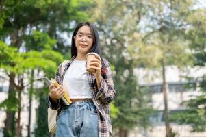 un hembra estudiante que lleva su mochila, digital tableta, y un café taza en pie en un instalaciones parque. foto