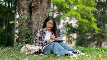 A young Asian woman sits under a tree in a campus park, holding a coffee cup and a tablet. photo