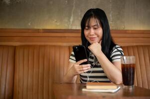 un asiático mujer es escuchando a música en su auriculares y utilizando su teléfono inteligente en un café tienda. foto