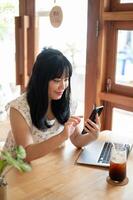 A happy Asian woman sits in a beautiful comfortable coffee shop, using her smartphone. photo