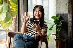 A smiling young Asian woman is sitting by the window in a vintage cafe with a smartphone in her hand photo