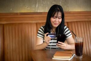 un mujer participación un teléfono inteligente y un crédito tarjeta a un mesa en un café comercio, pago cuentas en línea foto