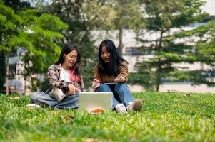 Two young Asian female college students are sitting on the grass in a park, using a laptop together. photo