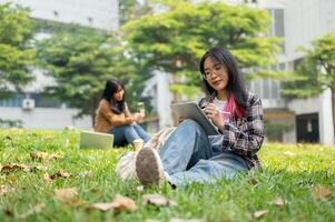 un hembra Universidad estudiante es estudiando en su digital tableta mientras sentado en césped en un parque. foto